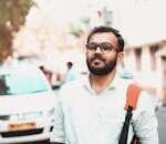 Confident man with glasses and backpack on a sunny urban street with cars in the background.